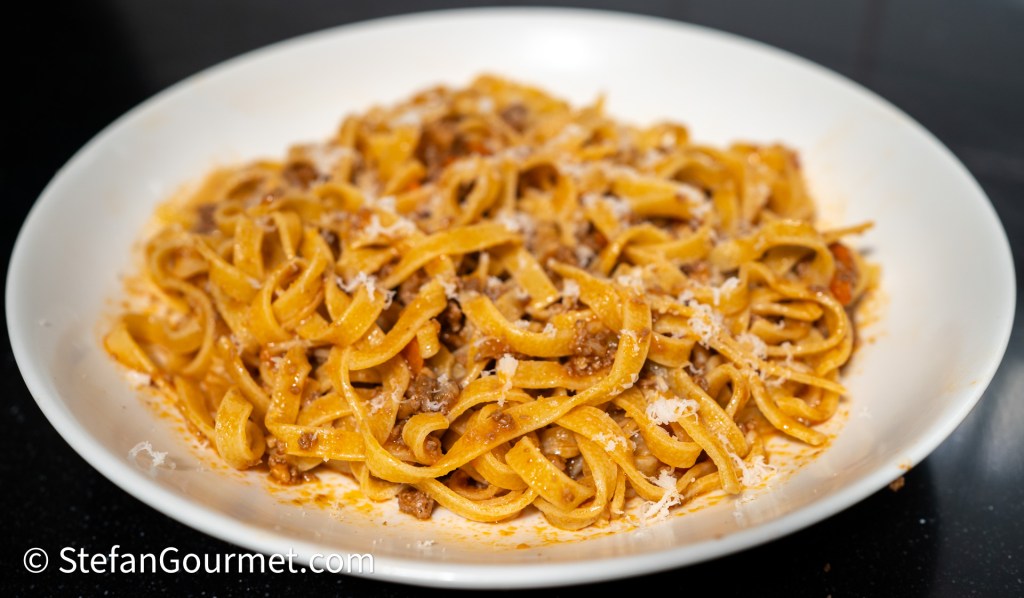 A serving of tagliatelle pasta with Bolognese sauce, garnished with grated cheese, presented in a white bowl against a dark background.