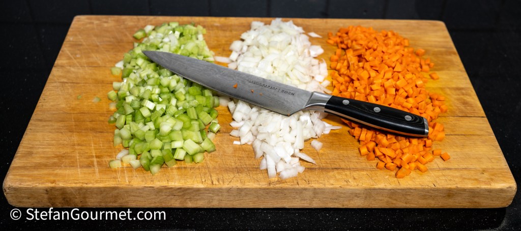 Chopped celery, onion, and carrot on a wooden cutting board with a knife.