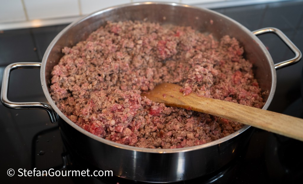 A pot filled with coarsely ground beef being sautéed, with a wooden spatula stirring the meat.
