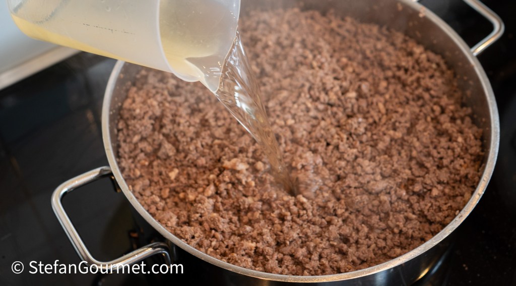 Pouring liquid into a pan with coarsely ground beef cooking on the stove.