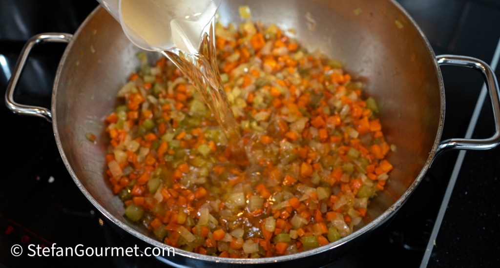 Pouring white wine over sautéed diced vegetables in a large stainless steel pan.