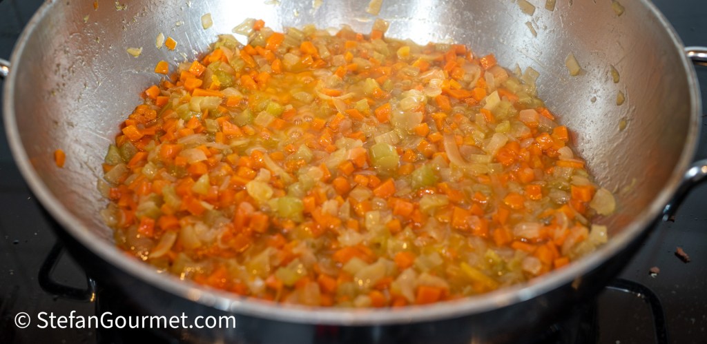 A sautéed mixture of finely diced carrots, celery, and onions simmering in a stainless steel pan.