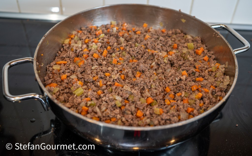 A large stainless steel pan containing cooked coarsely ground beef mixed with finely diced carrots, celery, and onions.