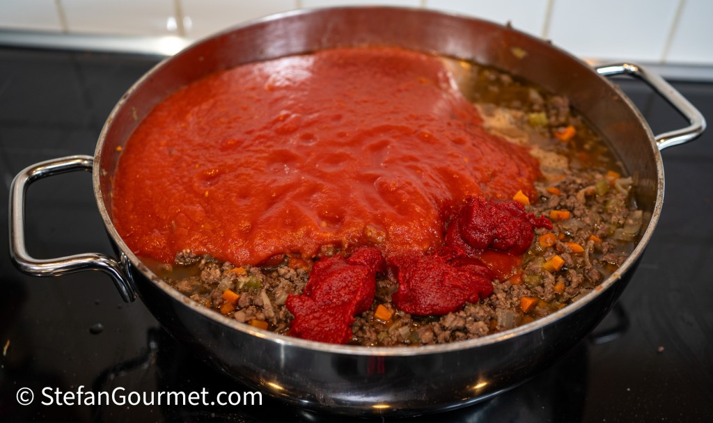 A large cooking pot filled with coarsely ground beef, diced vegetables, and a layer of tomato sauce and tomato paste for making Ragù alla Bolognese.