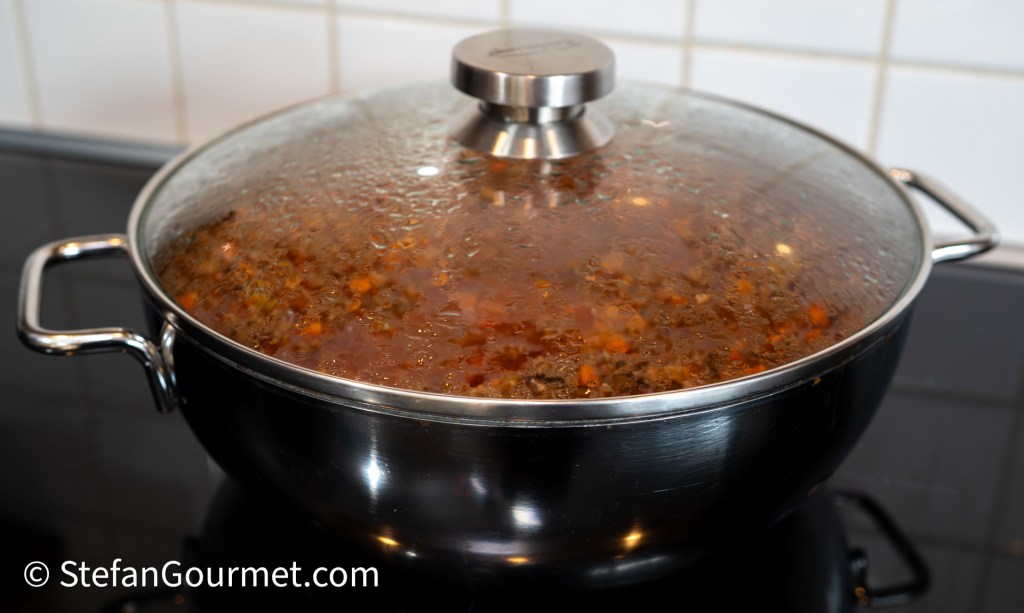 A pot simmering with Bolognese sauce covered by a lid, displaying a rich, steamy mixture with visible ingredients.
