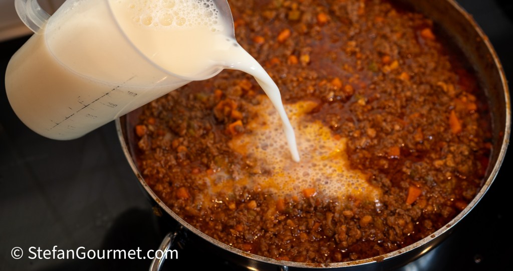 Pouring whole milk into a simmering pan of Bolognese sauce containing ground meat and vegetables.