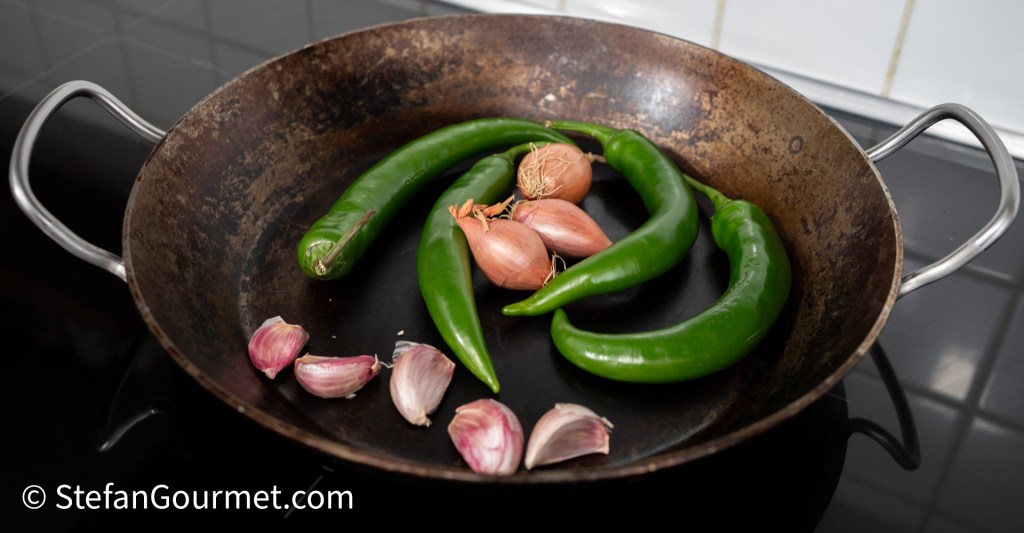 A frying pan filled with green chili peppers, shallots, and garlic, arranged on a dark surface.