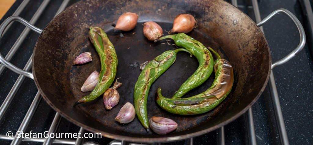 A frying pan with charred green chili peppers and whole shallots on a stove.