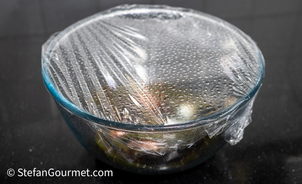 A glass bowl covered with plastic wrap, containing an assortment of ingredients for a Thai dip, sitting on a black countertop.