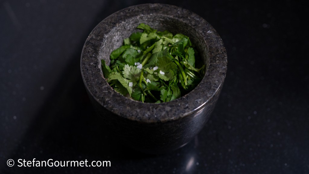 A stone mortar filled with fresh cilantro and a pinch of coarse salt, set against a dark background.