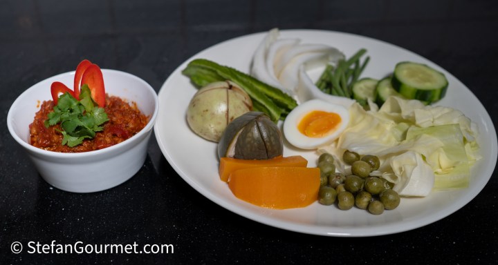 A plate featuring an assortment of fresh vegetables including cucumber, long beans, and cabbage, alongside a bowl of Nam Phrik Ong dip garnished with fresh herbs and chili slices.