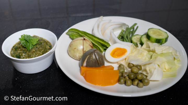 A plate featuring Nam Phrik Num, a green chili dip, accompanied by an array of raw and steamed vegetables, hard-boiled eggs, and various side items.