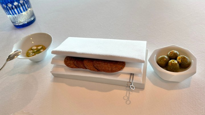 A white table setting featuring crispy crackers stacked on a napkin holder, accompanied by a small dish of olives and a bowl of green olives.