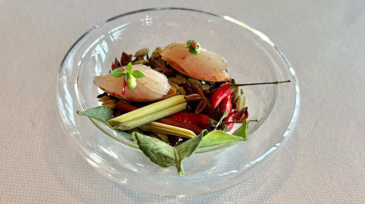 A glass bowl containing raw hamachi slices on a bed of spices and garnishes, including lemongrass, red chilies, and herbs.