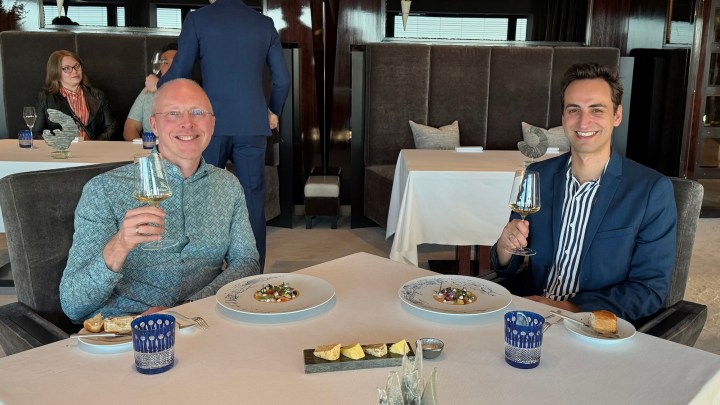 Two men seated at a table in a fine dining restaurant, smiling and holding wine glasses. A table set with gourmet dishes, including intricate plates of food, bread, and small condiments. Elegant restaurant interior in the background.