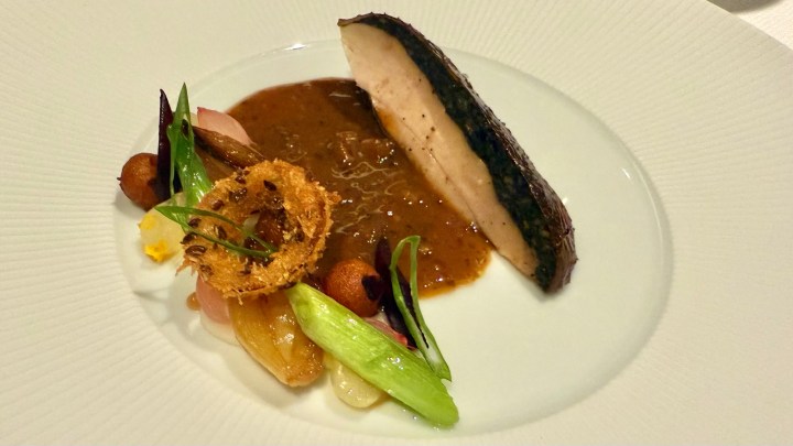 A beautifully plated dish featuring a piece of chicken breast with a crispy skin served alongside a rich sauce, accompanied by various vegetables and garnishes on a white plate.