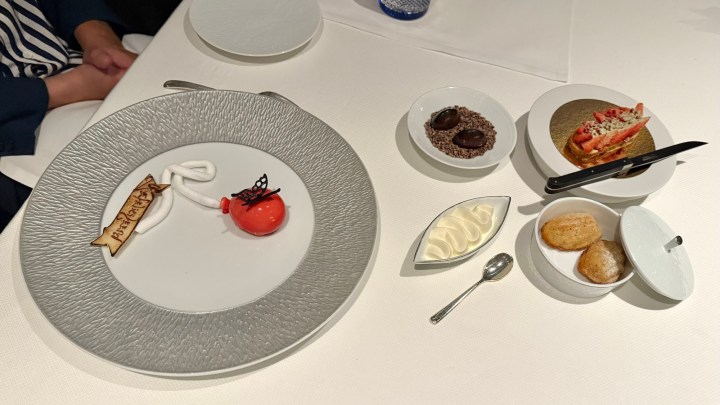 A beautifully plated dessert with a red dome, a chocolate decoration, and a personalized message on a wafer cookie, alongside a selection of additional sweet treats on a white tablecloth.