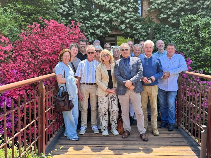 A group of wine connoisseurs posing together on a wooden bridge surrounded by vibrant pink and green flowers.