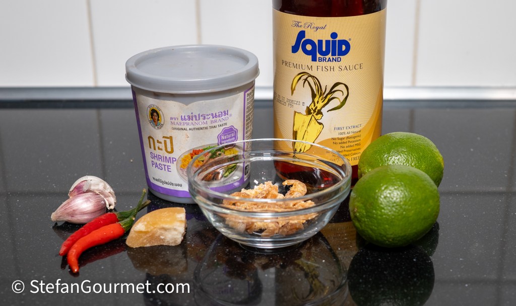 Ingredients for Nam Phrik Kapi dip, including shrimp paste, fish sauce, garlic, chilies, palm sugar, and limes, arranged on a black countertop.
