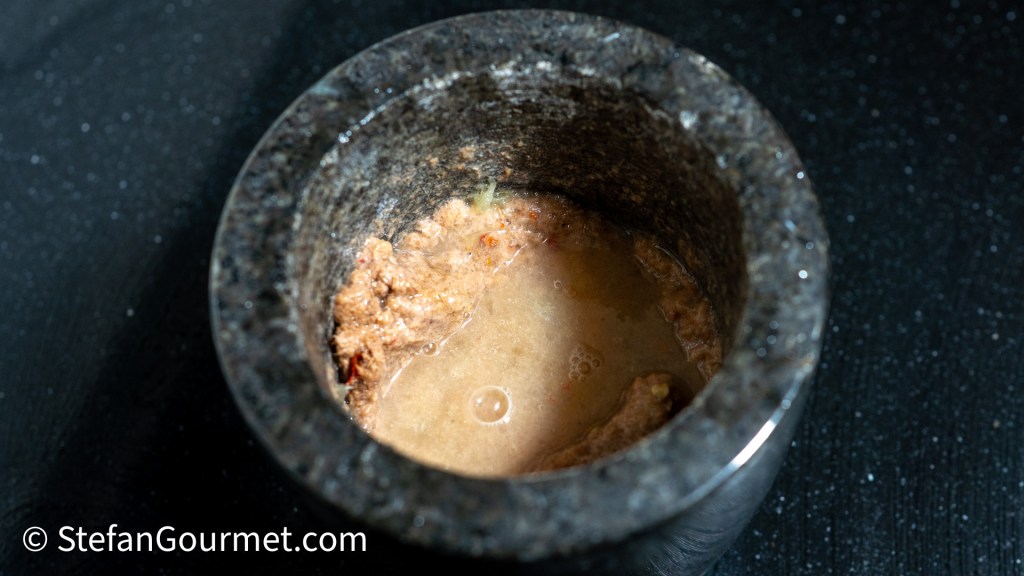 A mortar filled with a mixture of Nam Phrik Kapi dip, showcasing a textured blend of ingredients, set against a dark surface.
