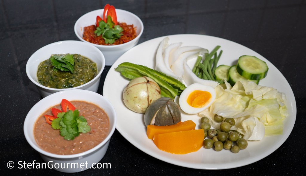 A plate featuring an assortment of Northern Thai hors d'oeuvres, including three bowls of dips and various fresh vegetables such as cucumber, cabbage, and green beans, along with hard-boiled eggs and slices of pumpkin.