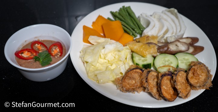 A plate of Northern Thai hors d'oeuvres featuring an assortment of raw and steamed vegetables, deep-fried eggplants, hard-boiled eggs, and a bowl of Nam Phrik Kapi dip garnished with red chilies and cilantro.