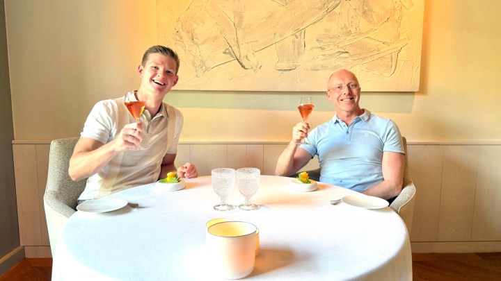 Two men sitting at an elegantly set table in a restaurant, raising glasses filled with cocktails in a toast, with an abstract artwork on the wall behind them.
