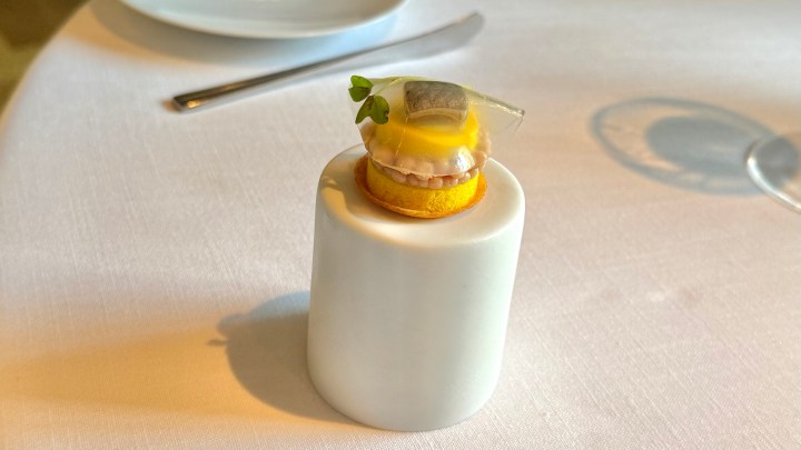 An elegant appetizer presented on a small white pedestal, featuring a delicate layer of fish topped with a translucent gel and a sprig of microgreens, all on a vibrant yellow base.