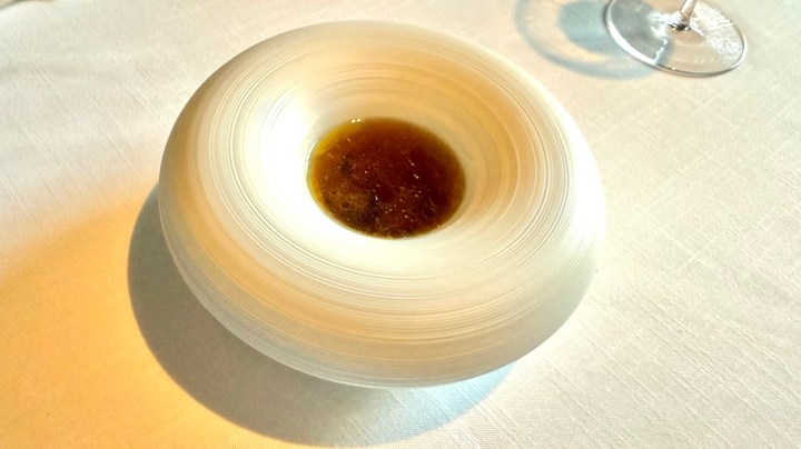 A minimalist dish served in a sleek white bowl, featuring a dark broth in the center, set against a plain tablecloth.