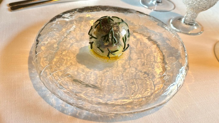 A clear glass plate holding a spherical dish decorated with edible leaves and a yellow base, presented elegantly on a white tablecloth.