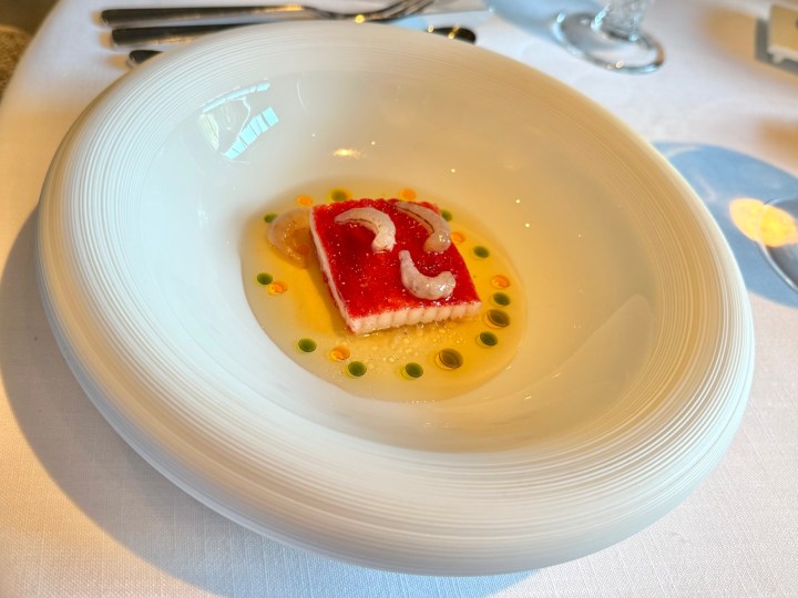 A beautifully plated dish featuring a rectangular piece of translucent seafood, topped with red sauce and garnished with small shrimp, served in a bowl with colorful sauce dots.