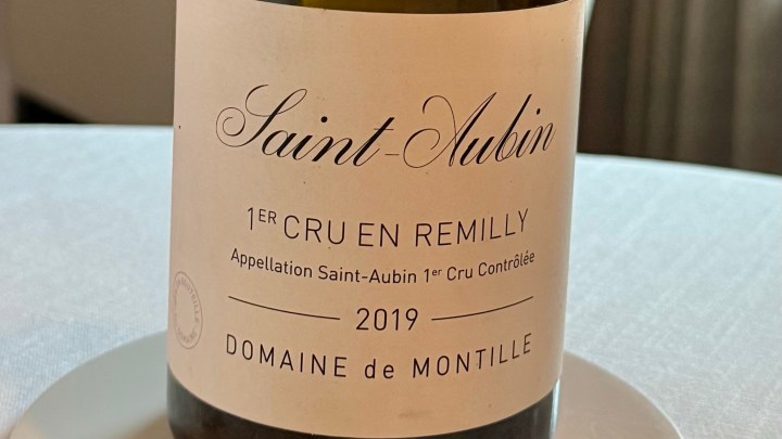 A close-up of a wine bottle labeled 'Saint-Aubin 1er Cru En Remilly 2019' from Domaine de Montille, placed on a white tablecloth.