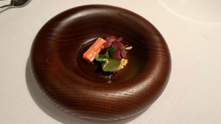 An elegant dish presented in a round wooden plate featuring small pieces of rhubarb, green sauce, and garnished with herbs.