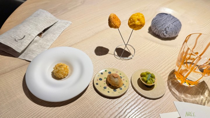 A beautifully arranged selection of snacks on a wooden table, featuring a white plate with a round crispy item, two colorful snacks on skewers, and small dishes containing various bites, alongside a ball of wool and a glass.