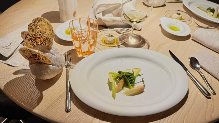 A beautifully set dining table with a minimalist white plate holding a delicate dish of green vegetables, surrounded by elegant glassware and artisanal bread.