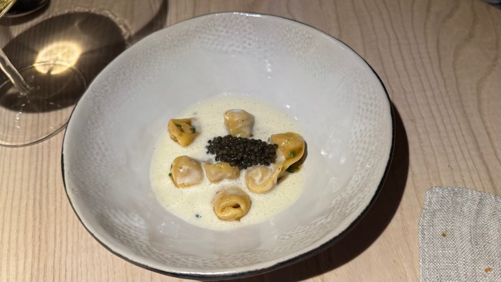A close-up of a bowl containing tortellini in a creamy sauce, topped with caviar, on a wooden table.