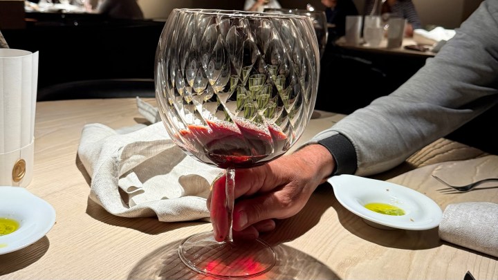 Close-up of a large, ornate wine glass filled with red wine, held by a person's hand on a wooden table.