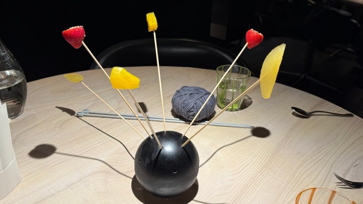 A decorative black sphere holding skewers with pieces of marinated fruit, including strawberries and tropical fruit, on a wooden table alongside a ball of wool and glassware.