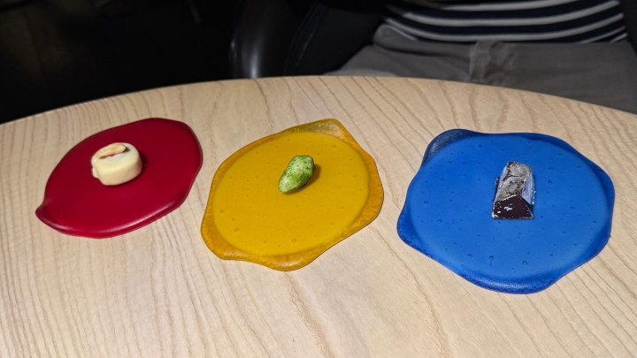 Three colorful dessert components on a wooden table, featuring a red piece, a yellow piece with a green item, and a blue piece with a chocolate square.