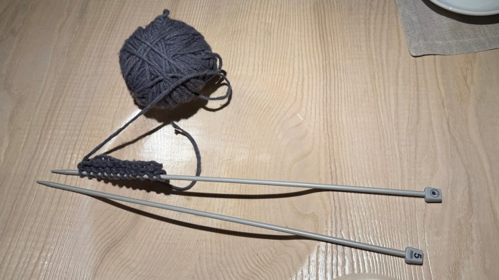 A ball of gray wool and knitting needles on a wooden table, with a small section of knitted fabric.