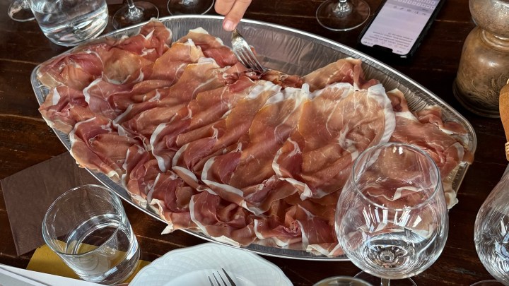 A large silver tray filled with thinly sliced prosciutto, accompanied by wine glasses and a dining setting.