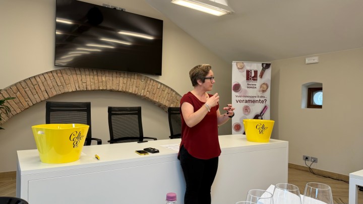 A woman giving a presentation in a meeting room, standing in front of a white table with yellow bowls and wine glasses set up, with a large screen in the background displaying a wine-related banner.
