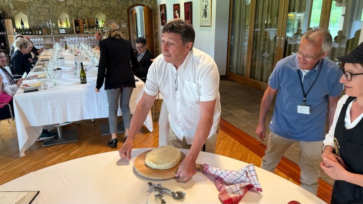 A group of people at a dining table, with one man presenting a round loaf of bread in the foreground, while others are seated and chatting in the background.