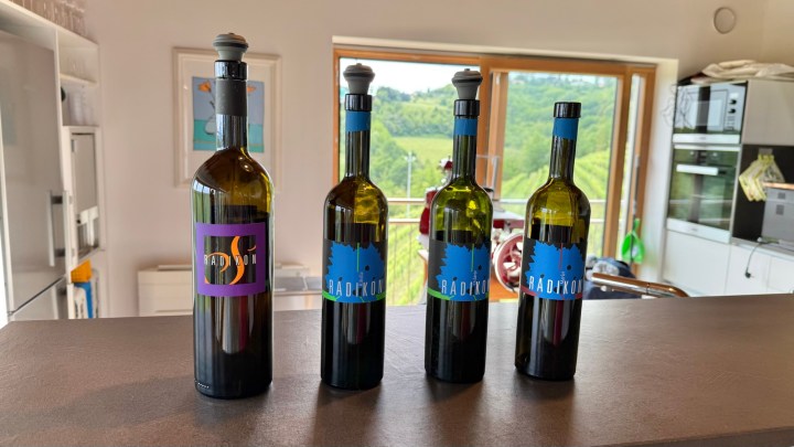 Four bottles of Radikon wine displayed on a countertop, with a scenic view of green hills visible through a window in the background.