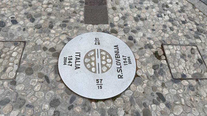 A round metal plaque on a cobblestone path indicating the border between Italy and Slovenia, with the years 1947 and 2004 engraved.