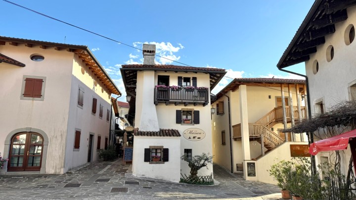 A picturesque view of a medieval village in Brda, Slovenia, featuring traditional architecture with two-story buildings, balconies adorned with flowers, and a clear blue sky overhead.