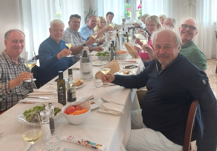 A group of people seated at a long table, raising glasses in a toast during a lunch gathering, surrounded by plates of food and wine bottles.