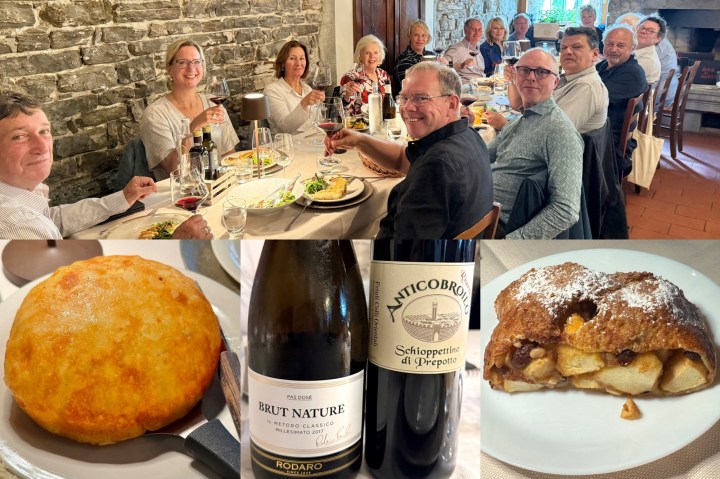 Group of diners enjoying a meal at a restaurant, featuring plates of food and wine bottles. In the foreground, a golden potato and cheese pancake, a bottle of Rodaro Brut Nature sparkling wine, and a bottle of Anticobroilo Schioppettino di Prepotto wine. Background shows diners engaged in conversation, with natural light coming through a window.