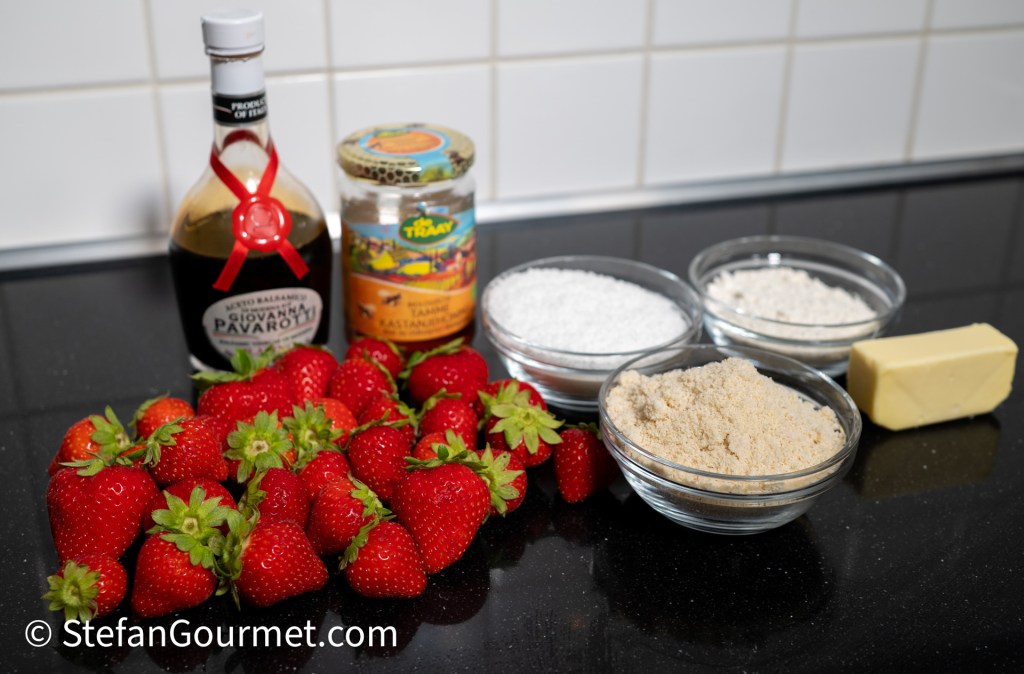 A selection of ingredients for a dessert, including fresh strawberries, a bottle of balsamic vinegar, almond flour, granulated sugar, and butter, arranged on a dark countertop.