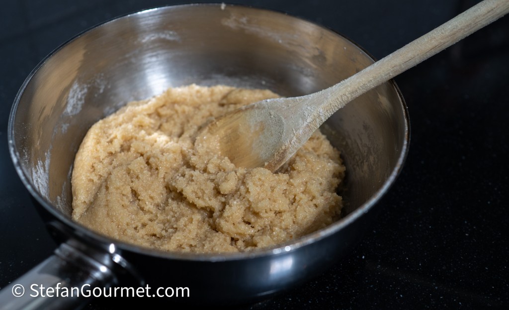 A mixing bowl containing a creamy almond dough with a wooden spoon resting inside, set against a dark background.